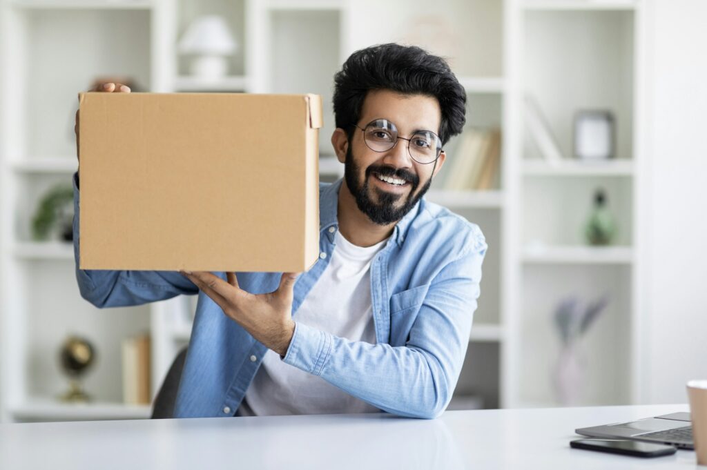 Delivery Concept. Happy Indian Man Demonstrating Big Cardboard Box At Camera